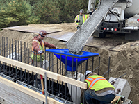 Workers assist cement truck pour of cement through a funnel to abutment base.